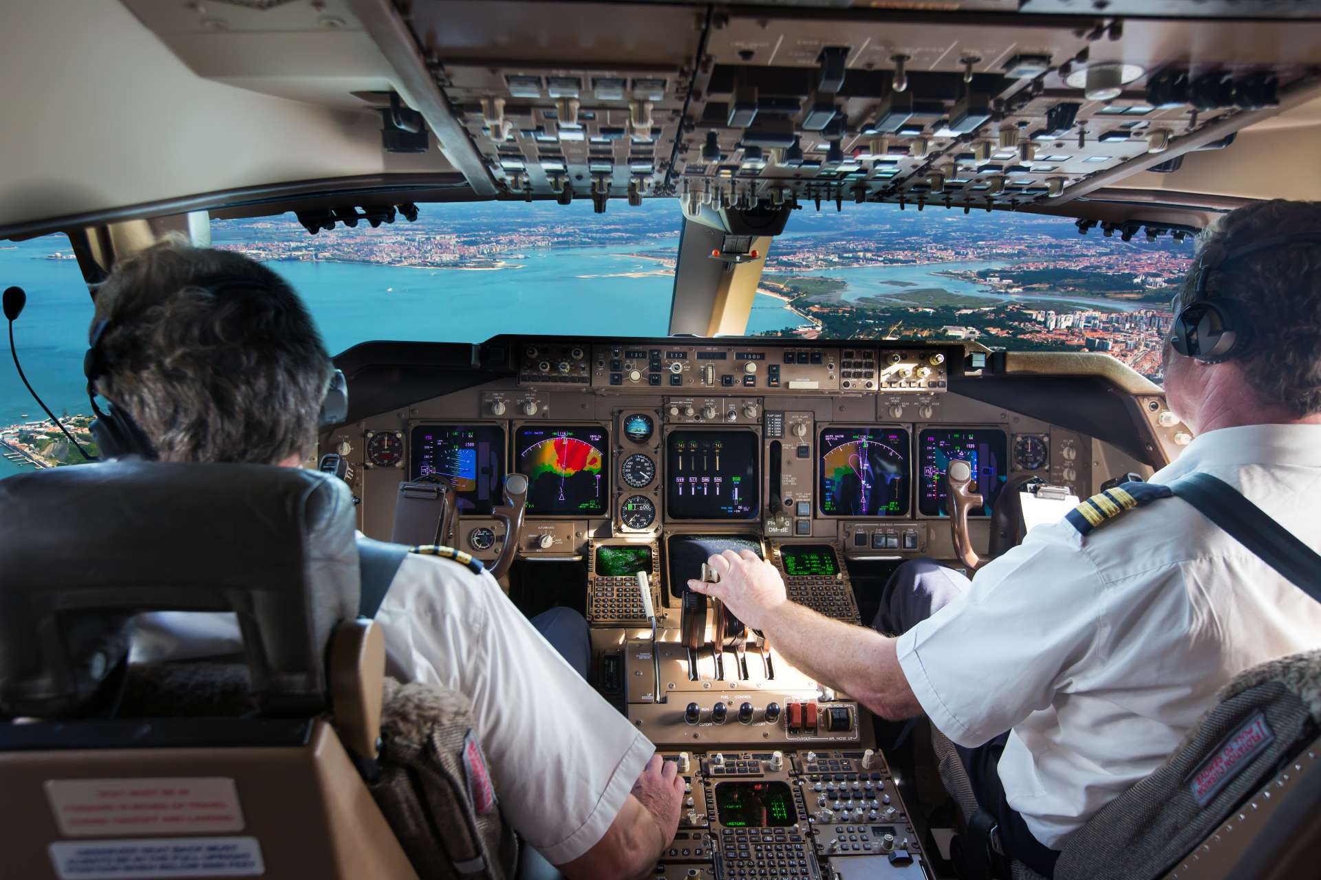 Two CRM airline pilots operating aircraft cockpit controls in flight, cityscape visible ahead