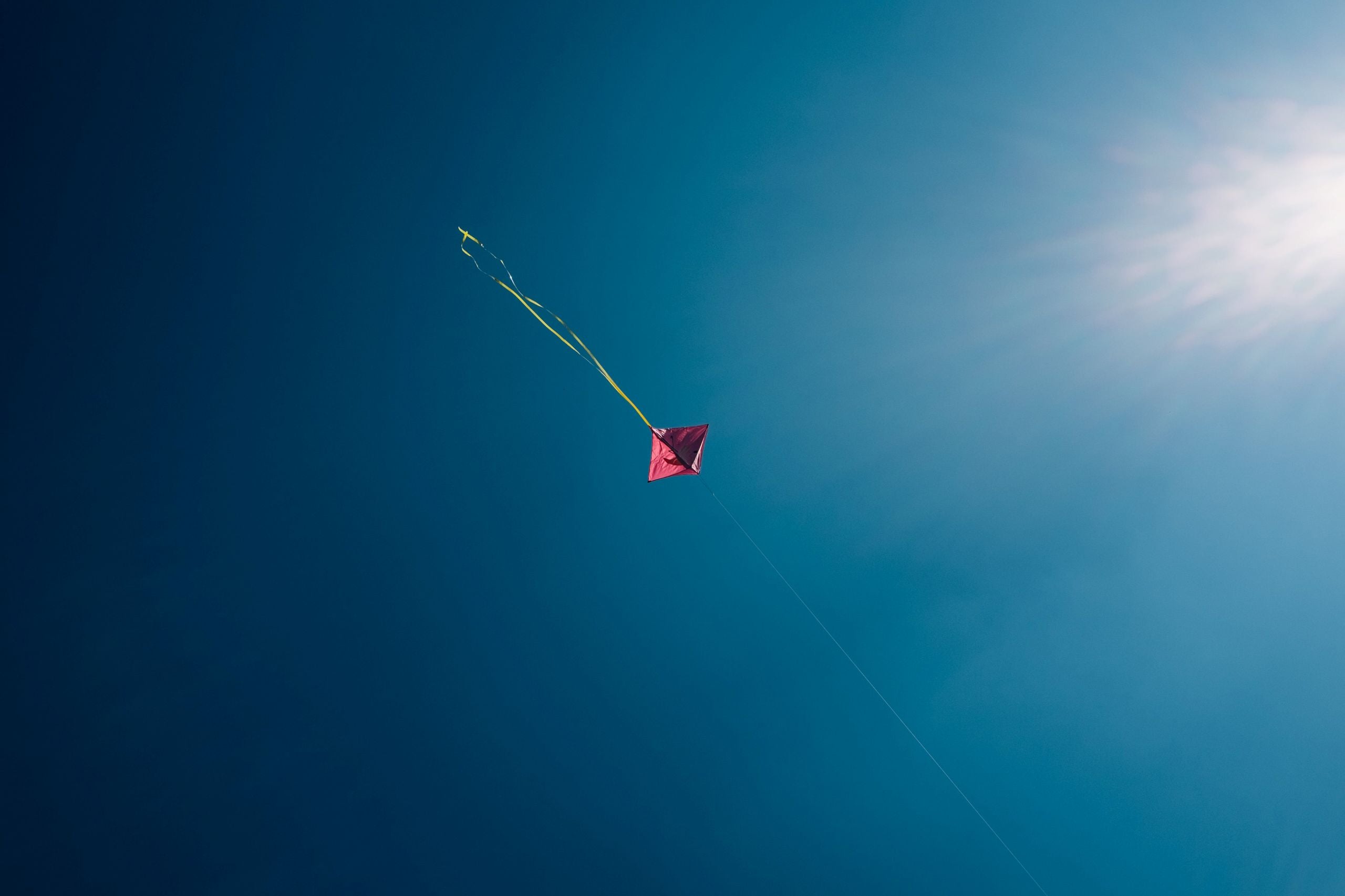 Minimalist pink kite with yellow tail flying in clear blue sky, sunlight shining.